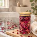 Glass jar with wooden lid containing dried rose petals on a wooden board by a bathtub.