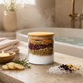 Glass jar with layered herbs and a wooden lid on a bathroom counter with a bathtub in the background.